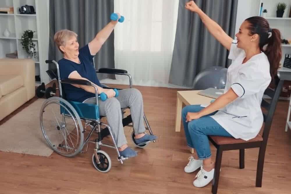 fisioterapeuta ayudando anciana silla ruedas a hacer ejercicios en casa